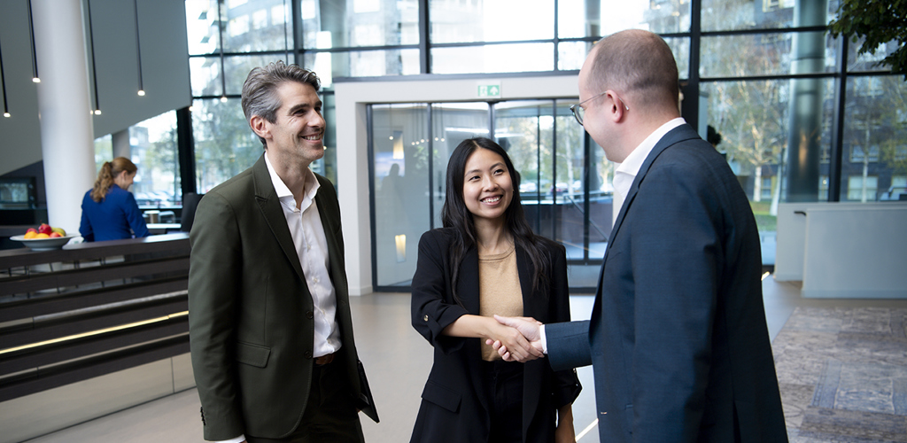 Three colleagues shake hands while smiling, in the lobby of an office building