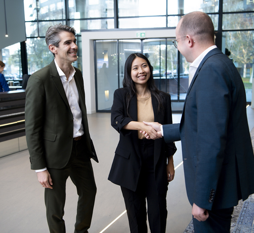 Three colleagues shake hands while smiling, in the lobby of an office building